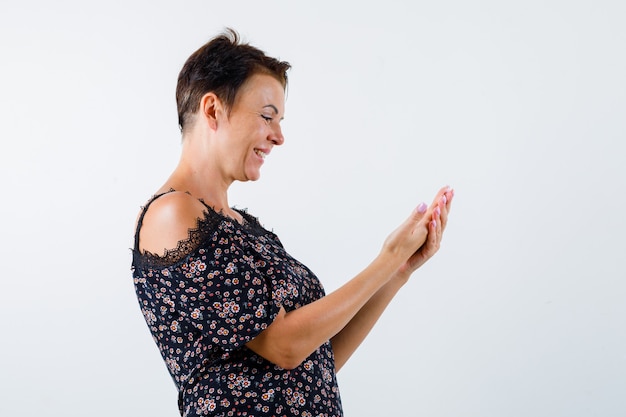 Rijpe vrouw in bloemenblouse, zwarte rok die hand uitrekt als iets vasthoudt, glimlachend en op zoek vrolijk, vooraanzicht.