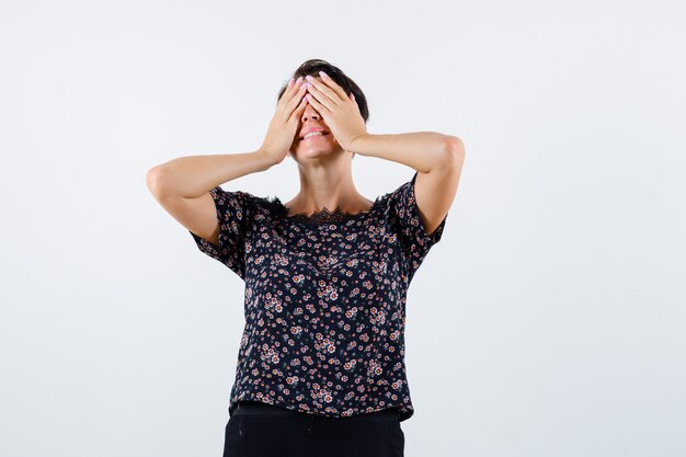 Rijpe vrouw in bloemenblouse, zwarte rok die ogen met handen bedekt en vrolijk, vooraanzicht kijkt.