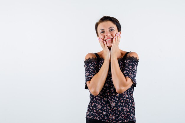 Rijpe vrouw in bloemenblouse, zwarte rok hand in hand op wangen, lachend en kijkend vrolijk, vooraanzicht.