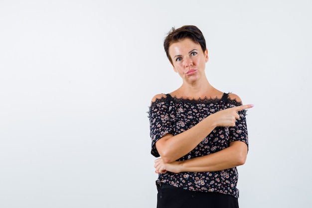 Rijpe vrouw in blouse die naar de rechterkant wijst en zelfverzekerd, vooraanzicht kijkt.