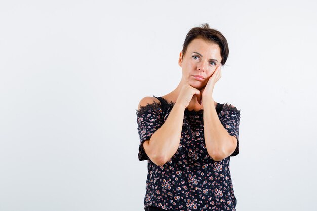 Rijpe vrouw in gebloemde blouse, zwarte rok, wang op handpalm leunend, kin aan kant steunen, aan iets denken en peinzend, vooraanzicht kijken.