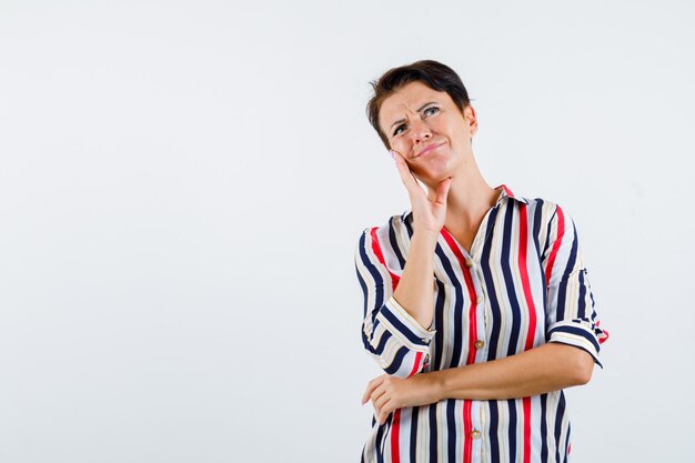 Rijpe vrouw in gestreepte blouse die wang op palm leunt terwijl aan iets denkt, hand op taille houdt en peinzend, vooraanzicht kijkt.