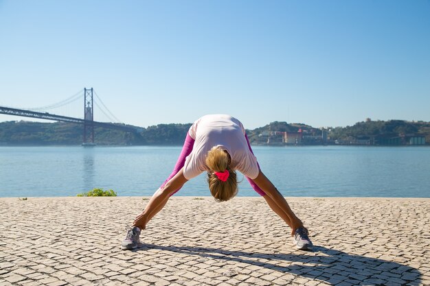 Rijpe vrouw in sportkleren die ochtendoefening buiten doen, rug en benen spieren strekken bij rivier. Activiteit of actief levensstijlconcept
