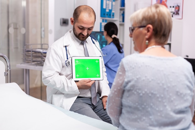 Rijpe vrouw in ziekenhuis spreekkamer luisteren arts met tablet groen scherm