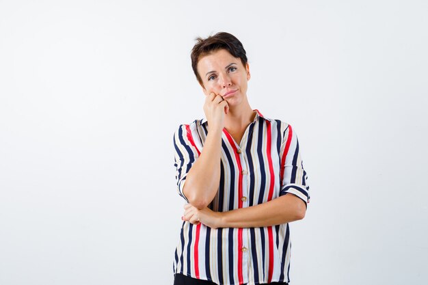 Rijpe vrouw leunende wang op handpalm, houdt een hand onder de elleboog in een gestreept overhemd en kijkt peinzend. vooraanzicht.