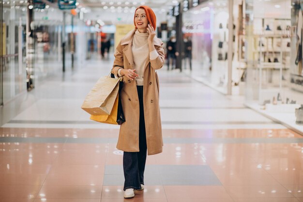Rijpe vrouw met boodschappentassen in winkelcentrum met behulp van telefoon