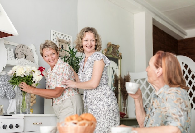 Rijpe vrouw met dochter die bloemenvaas op keukenteller schikken terwijl haar moeder die koffie hebben