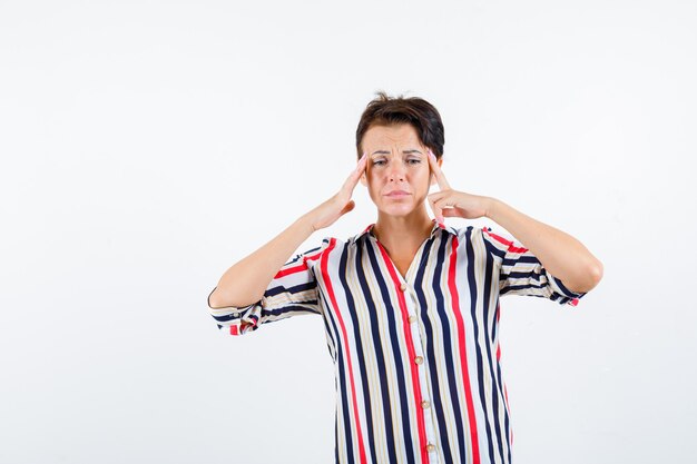 Rijpe vrouw met wijsvingers op tempels in gestreepte blouse en op zoek naar somber, vooraanzicht.