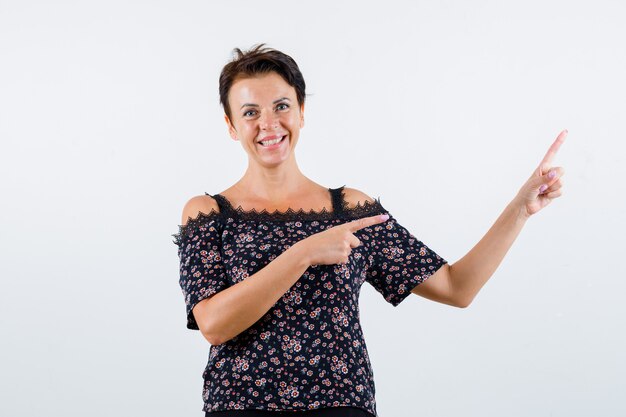 Rijpe vrouw naar rechts wijzend met wijsvingers in bloemenblouse, zwarte rok en op zoek gelukkig, vooraanzicht.