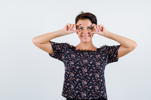 Rijpe vrouw ogen openen met vingers in bloemenblouse, zwarte rok en vrolijk kijken. vooraanzicht.