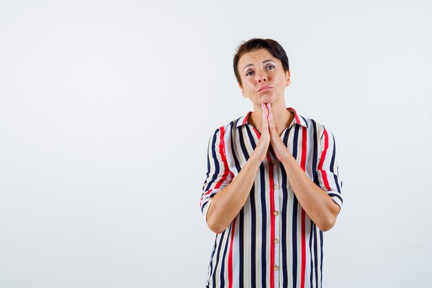 Rijpe vrouw omklemt de handen in gebedspositie in gestreept overhemd en kijkt geconcentreerd. vooraanzicht.