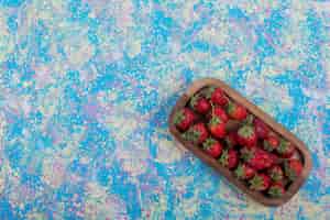 Gratis foto rode aardbeien in een houten schotel op de blauwe tafel, bovenaanzicht