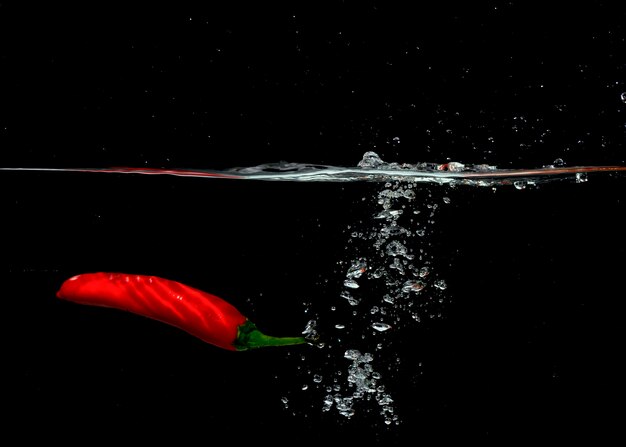 Rode Spaanse peper die met bellen in water tegen zwarte achtergrond valt