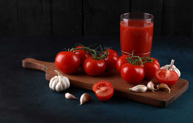 Rode tomaten en knoflookhandschoenen op de houten raad met een glas sap op de blauwe achtergrond.