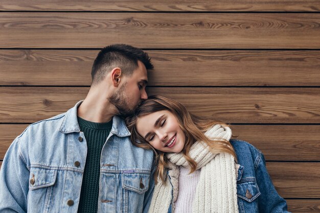 Romantisch blond meisje liggend op de schouder van vriendje met gesloten ogen. Indoor portret van Europese brunette man kussen vriendin haar op houten muur.