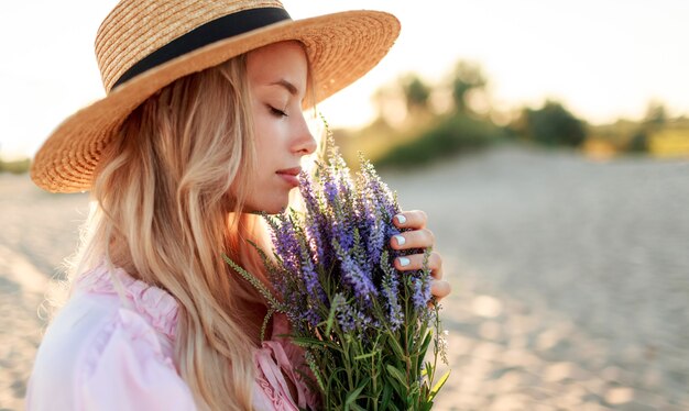 Romantisch close-up portret o charmant blond meisje in strohoed ruikt bloemen op het avondstrand, warme zonsondergangkleuren. Boeket van lavendel. Details.