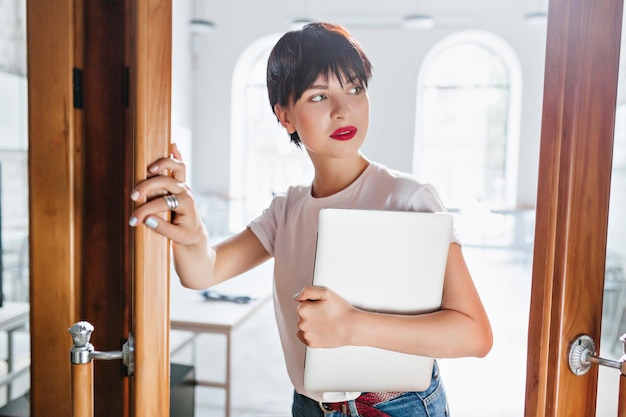 Romantisch donkerharig meisje met laptop die wegkijkt, wachtend op iemand naast de deur. Indoor portret van jonge vrouw in informele kleding poseren met computer met witte kantoorruimte op de achtergrond.