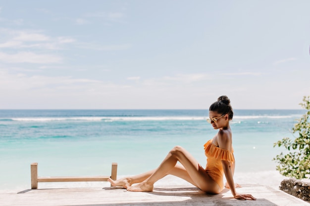 Romantisch gelooid meisje in oranje kledij zittend op het strand. Betoverend wit vrouwelijk model in zonnebril poseren op de grond op Zeekust.