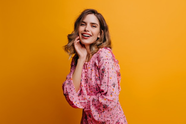 Romantisch meisje in roze blouse poseren met oprechte glimlach. Binnenfoto van vrij krullende vrouw die pret op gele muur heeft.