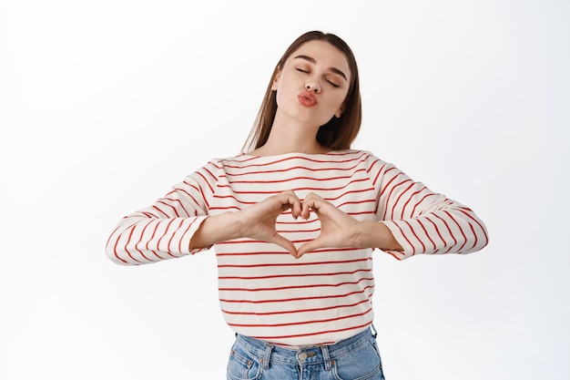 Romantisch meisje toont hart, ik hou van je, teken en zoen, lippen tuit voor luchtkus, druk tederheid en genegenheid uit, sympathie, zoals iemand, staande over een witte muur