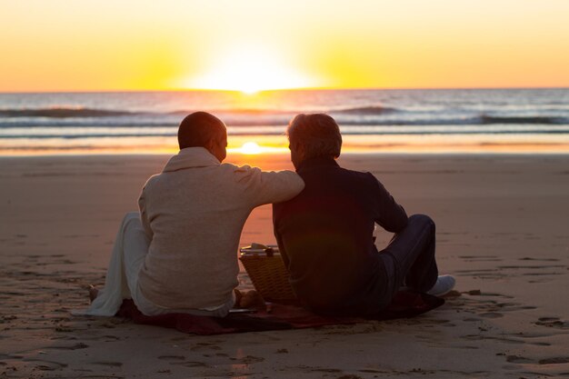 Romantisch senior paar met picknick bij zonsondergang op het strand. Man en vrouw zitten op deken aan de kust en genieten van een fantastisch uitzicht op het zeegezicht. Achteraanzicht. Romantiek, lifestyle, vrijetijdsconcept