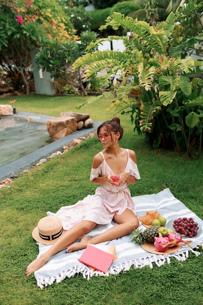 Romantisch sierlijke Aziatische model zittend op deken, wijn drinken en genieten van zomerpicknick in tropische tuin.