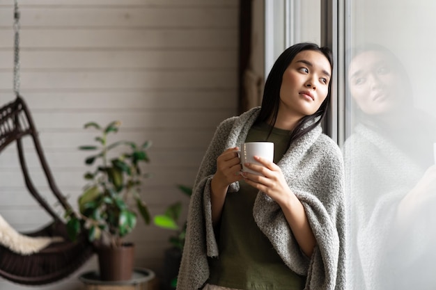 Romantische aziatische vrouw gewikkeld in een deken, leunend op het raam en kijkend naar buiten terwijl ze hete koffie drinkt