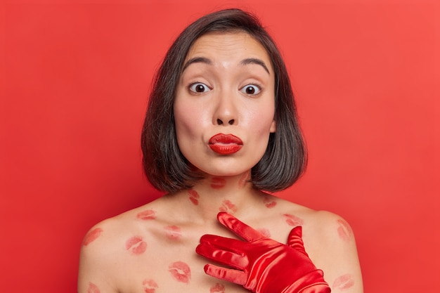 Romantische Aziatische vrouw stuurt luchtkus naar camera houdt rood geschilderde lippen afgerond heeft tedere uitdrukking poses met naakte lichaam gezonde huid draagt elegante lange handschoenen