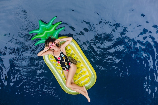 Gratis foto romantische jonge vrouw rusten in zwembad in warme zomerdag. overhead portret van blije europese dame in zonnebril en zwembroek.