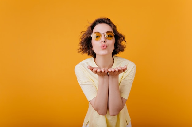 Romantische kortharige vrouw in vintage glazen poseren met mooie gezichtsuitdrukking. Blij meisje in geel t-shirt luchtkus verzenden tijdens fotoshoot.