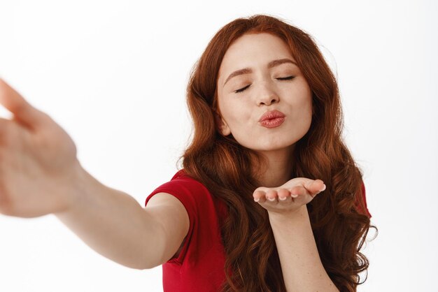 Romantische roodharige vrouw die luchtkus verzendt met schattig flirterig gezicht dat selfie of videobellende minnaar flirterig en koket vaarwel staande tegen een witte achtergrond