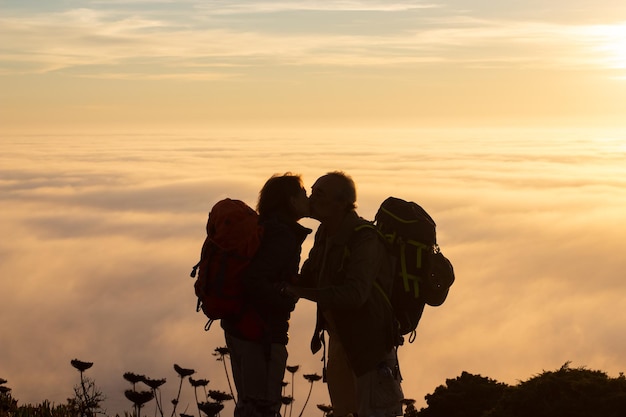 Romantische silhouet wandelaars bij zonsondergang. Man en vrouw in vrijetijdskleding en met munitie staan op het hoogtepunt, zoenen. Hobby, actieve levensstijl, liefdesconcept