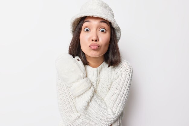 Romantische tedere brunette Aziatische vrouw houdt lippen gevouwen wil je kussen draagt witte bontmuts gebreide trui en wanten geniet van wintertijd geïsoleerd op witte achtergrond. Menselijke gezichtsuitdrukkingen