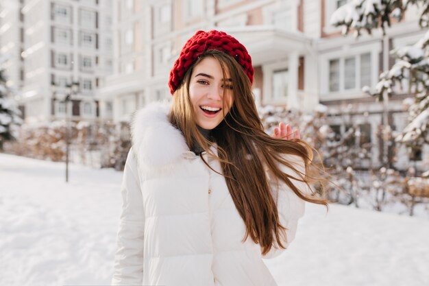 Romantische vrouw in rode gebreide muts speelt met haar lange bruine haar op besneeuwde straat. Buitenfoto van enthousiast Europees vrouwelijk model dat in de wintervakantie rondloopt.