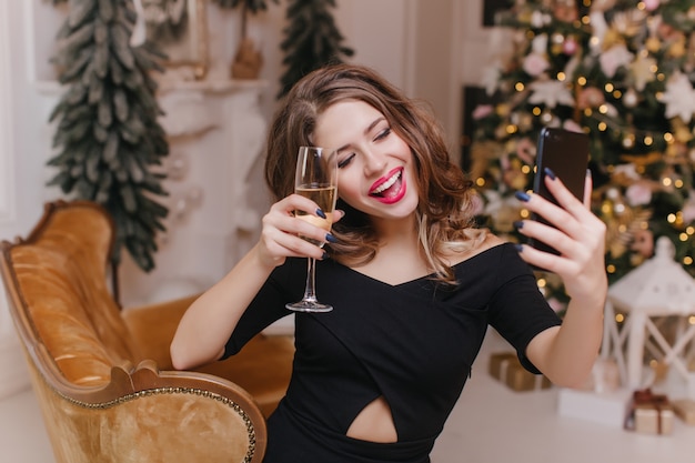 Romantische vrouw in stijlvolle zwarte kleding glas wijn verhogen en selfie maken. Emotionele dame die plezier heeft tijdens de wintervakantie en een foto van zichzelf maakt.