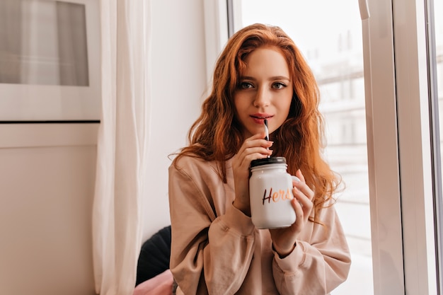 Romantische vrouw met golvend haar thuis cappuccino drinken. Indoor foto van schattige Europese dame chillen in haar kamer.