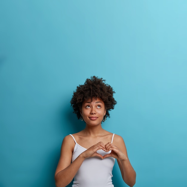 Romantische zorgzame tedere Afro-Amerikaanse vrouw drukt passie en romantiek uit, maakt hartgebaar, kijkt omhoog, gekleed in casual vest, geïsoleerd op blauwe muur, lege ruimte voor uw promotie