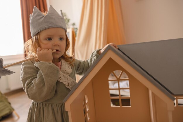 Roodharig klein kaukasisch meisje met speelgoedkroon en toverstaf in de buurt van poppenhuis thuis Jeugdconcept