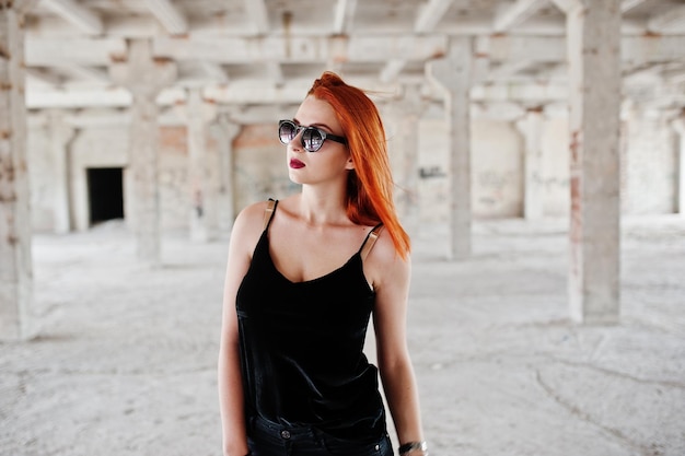 Gratis foto roodharig stijlvol meisje in zonnebril draagt in het zwart tegen verlaten plek