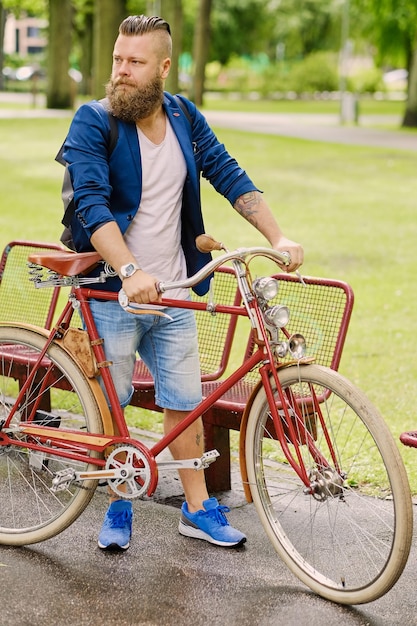 Roodharige bebaarde man gekleed in een blauwe jas en jeans op een retro fiets in een park.