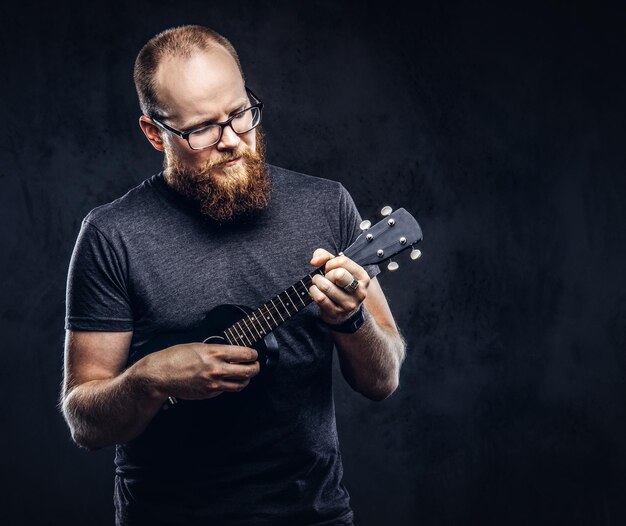 Roodharige, bebaarde mannelijke muzikant die een bril draagt, gekleed in een grijs t-shirt dat op een ukelele speelt. Geïsoleerd op donkere gestructureerde achtergrond.