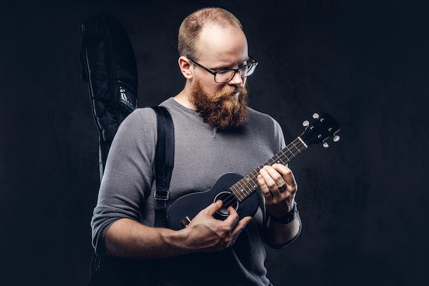 Roodharige, bebaarde mannelijke muzikant die een bril draagt, gekleed in een grijs t-shirt dat op een ukelele speelt. Geïsoleerd op donkere gestructureerde achtergrond.