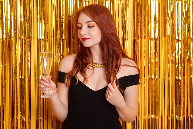 Roodharige dame met champagneglas, dromerig opzij kijkend, haar krulspelden aanraken, poseren tegen een gele muur versierd met glitter, meisje met zwarte jurk.