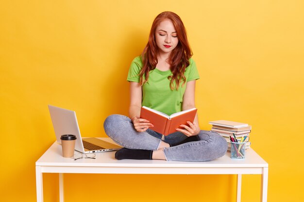 Roodharige geconcentreerde vrouw draagt groene t-shirt en spijkerbroek, houdt boeken in handen en leest, student zittend op tafel met gekruiste benen, dame omringd met laptop, pennen