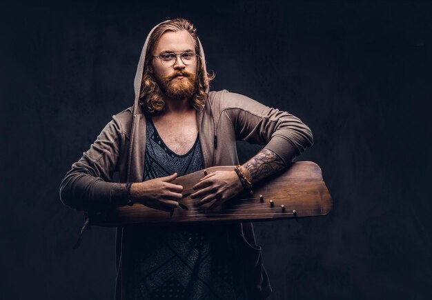 Roodharige hipster man met lang weelderig haar en volle baard gekleed in een hoodie en t-shirt spelen op een Russisch traditioneel muziekinstrument - gusli. Geïsoleerd op de donkere achtergrond.