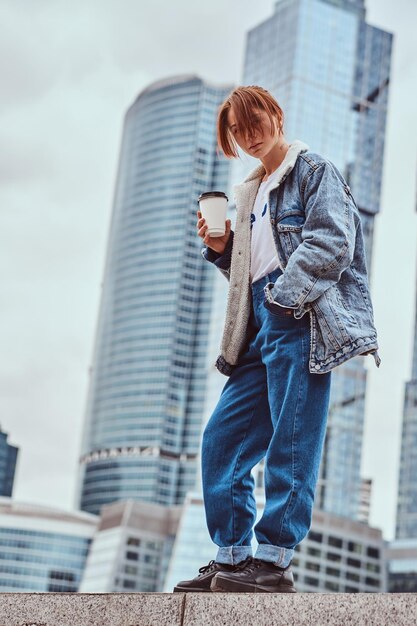 Roodharige hipster meisje met tatoeage op haar gezicht, gekleed in een denim jas met afhaalkoffie voor wolkenkrabbers in de stad Moskou.