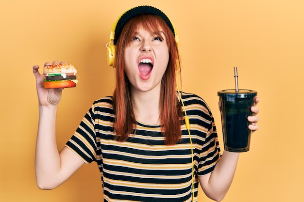 Roodharige jonge vrouw die een hamburger eet en frisdrank drinkt met een koptelefoon boos en gek schreeuwend gefrustreerd en woedend, schreeuwend van woede die omhoog kijkt.