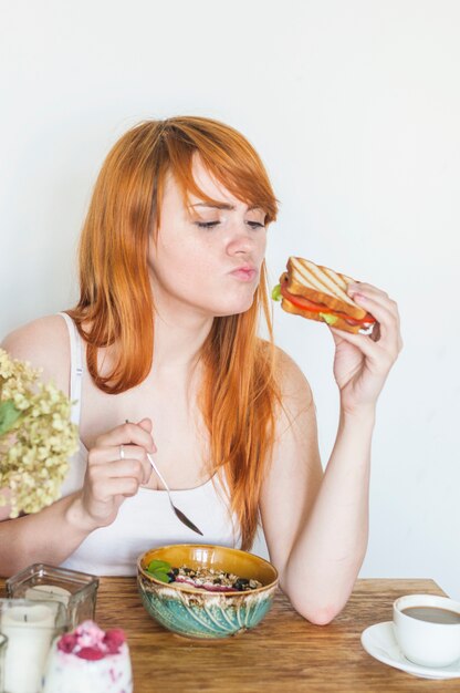 Roodharige jonge vrouw die sandwich met kom havermeel bekijkt