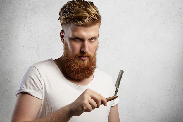 Gratis foto roodharige kapper of kapper met pluizige baard gekleed in een wit t-shirt met een scherp mes van zijn scheermes met rechte rand in de kapperszaak, klaar om zijn klanten te scheren.