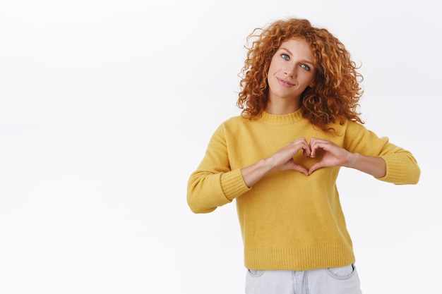 Gratis foto roodharige krullende vrouw in gele trui, bekent verliefd, toont hartteken boven borst